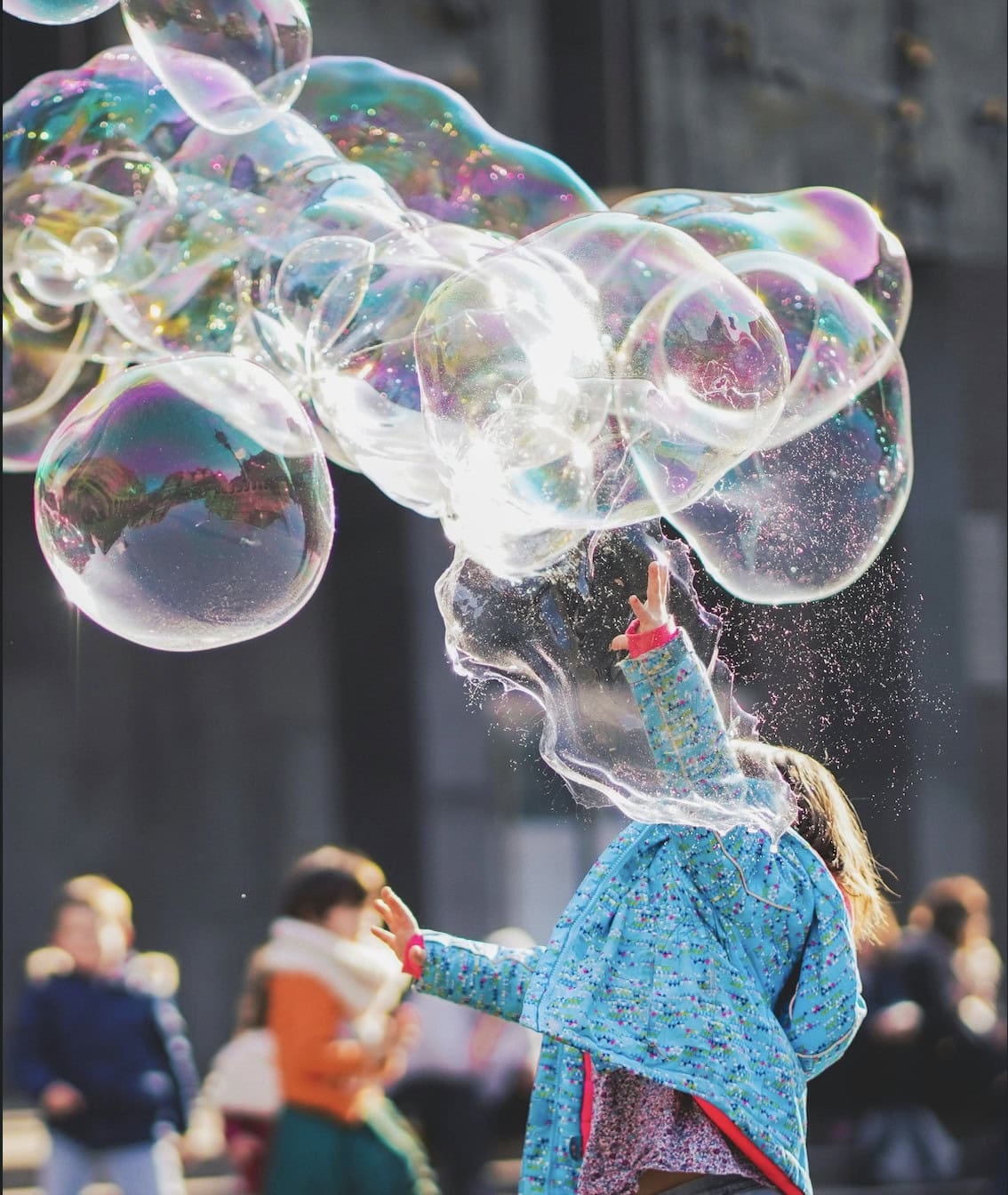 Child reaching up to pop giant soap bubbles in a sunny outdoor plaza.